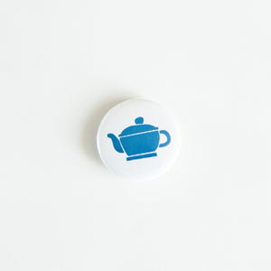 A button on white tabletop. The button features an illustration of a blue teapot. 