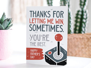 A greeting card featured standing up on a white tabletop with a pink plant pot in the background and some succulents in the pot. There’s a woven basket in the background with a cactus inside. The card features the words "Thanks for letting me win, sometimes. You're the best. Happy Father's Day." 