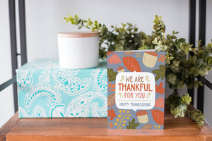 A greeting card is on a table top with a present in the background. There's greenery on top of the present. The card reads "We are Thankful for You, Happy Thanksgiving" with illustrated leaves and acorns around the words. 
