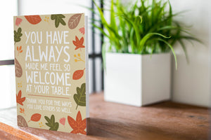 A photo of a card featured on a tabletop next to a white planter filled with a green plant. ​​The card features the words "You have always made me feel so welcome at your table. Thank you for the ways You love others so well" with illustrated leaves surrounding the words. 