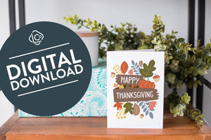 A greeting card is on a table top with a present in the background. There's greenery on top of the present. The card reads “Happy Thanksgiving” with illustrated leaves, a pumpkin, and acorn. The words "digital download" are featured in a circle over the image.