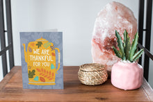 Load image into Gallery viewer, A greeting card featured standing up on a white tabletop with a pink plant pot with succulents, a pink rock and a small woven basket. The card features the words &quot;We are Thankful for You&quot; with the words featured inside an illustrated teapot.