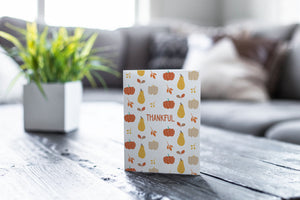 A greeting card featured on a black, wood coffee table. There’s a white planter in the background with a green plant. There’s also a gray sofa in the background with a white pillow. The card features the word "Thankful" with a pattern of illustrated pumpkins and leaves behind the word. 