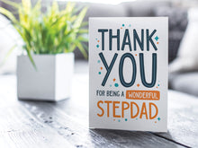 Load image into Gallery viewer, A greeting card featured on a black, wood coffee table. There’s a white planter in the background with a green plant. There’s also a gray sofa in the background with a white pillow. The card features the words “Thank You for Being a Wonderful Stepdad.”