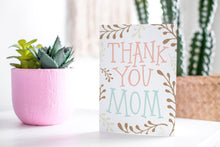 Load image into Gallery viewer, A greeting card featured standing up on a white tabletop with a pink plant pot in the background and some succulents in the pot. There’s a woven basket in the background with a cactus inside. The card features the words “Thank You Mom” with illustrated plant leaves.