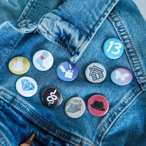 A photo of 10 pin badges on a jean jacket. The badges feature illustrations of a snake, a lighter, a cowboy boot,  cat, a butterfly, a house, a typewriter, a diamond, the number 13, and a black hat. 