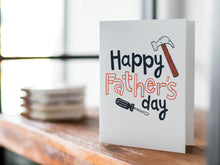 Load image into Gallery viewer, A card on a wood tabletop with an object in the background that is out of focus. The card features the words “Happy Father’s Day” with an illustrated hammer and screwdriver around the words. 