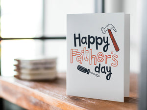 A card on a wood tabletop with an object in the background that is out of focus. The card features the words “Happy Father’s Day” with an illustrated hammer and screwdriver around the words. 