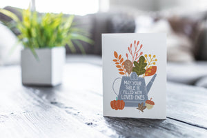 A greeting card featured on a black, wood coffee table. There’s a white planter in the background with a green plant. There’s also a gray sofa in the background with a white pillow. The card features the words "May Your Table be Filled with Loved Ones" with the words inside an illustrated watering can with leaves coming out of the top.