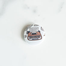 Load image into Gallery viewer, A buttons on a white tabletop. The button features an illustration of a typewriter.