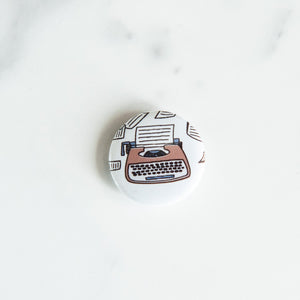 A buttons on a white tabletop. The button features an illustration of a typewriter.