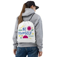 Load image into Gallery viewer, A woman carrying a white drawstring bag on her back. The drawstring bag has the words &quot;Be Yourself&quot; in blue with green illustrated sketchy pieces around the words. 