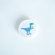 Load image into Gallery viewer, A button pin on a white tabletop. The button features an illustration of a Velociraptor. 