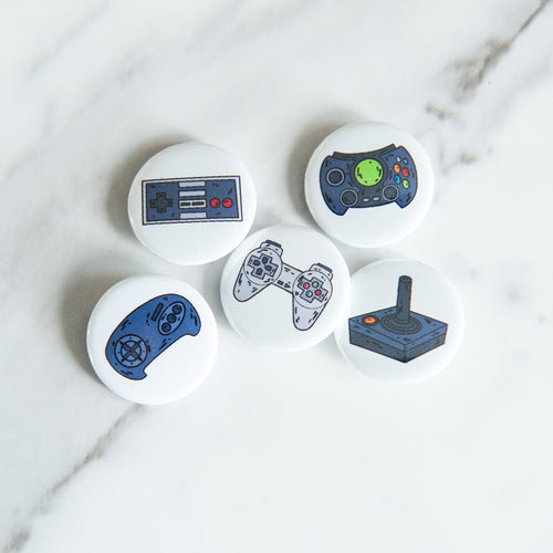 A set of five button pins on a white tabletop. The buttons feature five different illustrations of game controllers. 