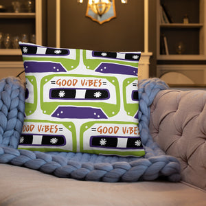 A white pillow on a sofa with a blue blanket. The white pillow features the artwork on a white background with an illustrated cassette tape pattern with the words 'good vibes.'