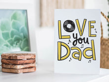 Load image into Gallery viewer, A greeting card featured standing up on a white tabletop with a framed photo of a succulent in the background and a stack of wooden coasters. There’s a woven basket in the background with a cactus inside. The card features the words “Love you Dad” with a vinyl record as the “O” of love and music notes around the letters. 