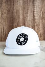 Load image into Gallery viewer, A white trucker hat on a tabletop. The hat features an black embroidered design with an illustrated vinyl record and the words &#39;good vibes&#39; inside.  