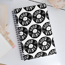 Load image into Gallery viewer, A spiral notebook on a white, wood tabletop. The white spiral notebook has black spiral binding. The pattern design features the words &quot;good vibes&quot; inside an illustrated vinyl record.  