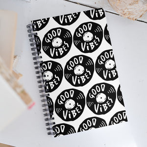 A spiral notebook on a white, wood tabletop. The white spiral notebook has black spiral binding. The pattern design features the words "good vibes" inside an illustrated vinyl record.  