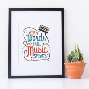 Artwork in a black frame with the with a white matte. The frame is leaning on a white counter with a terracota pot with a catcus next to it. The artwork features hand drawn lettering with the phrase "When words fail, music speaks." In the upper corner of the words an illustrated cassette tape is featured.