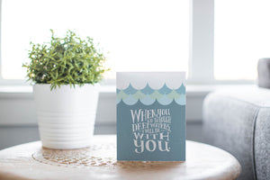 A greeting card laying on a wooden table with some cut wood details. The card features the words “When you go through deep waters, I will be with you.”