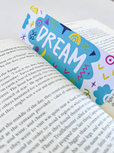 Load image into Gallery viewer, A bookmark placed inside a book. The bookmark design has the word &quot;dream&quot; inside a teal cloud-like shape with sketchy, whimsical items around. 