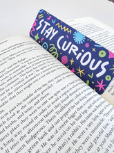 Load image into Gallery viewer, A bookmark placed inside a book. The bookmark has the words &quot;stay curious&quot; with fun, sketchy illustrations. 