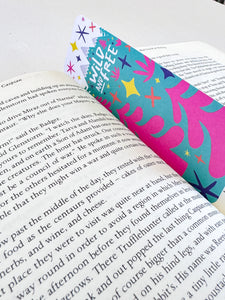 A bookmark placed inside a book. The bookmark features the words "wild and free" inside illustrated snow-capped mountains with illustrated trees. 