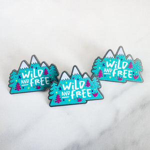 Three enamel pins featuring the words "wild and free" inside illustrated mountains with illustrated trees. 