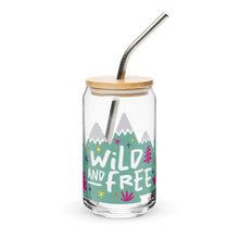 Load image into Gallery viewer, A photo of a glass can tumbler on a white background with a bamboo lid and silver metal straw. The design on the glass tumbler says the words &quot;wild and free&quot; inside illustrated mountains with sketchy trees around. 