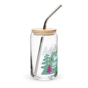 A photo of a glass can tumbler on a white background showing the side of the glass. The glass tumbler has a bamboo lid and silver metal straw. The design on the glass tumbler says the words "wild and free" inside illustrated mountains with sketchy trees around. 