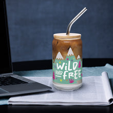 Load image into Gallery viewer, A photo of a glass can tumbler on a desktop next to a laptop. The glass tumbler has a bamboo lid and silver metal straw. The design on the glass tumbler says the words &quot;wild and free&quot; inside illustrated mountains with sketchy trees around. 