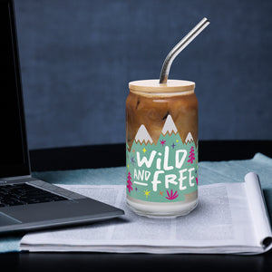 A photo of a glass can tumbler on a desktop next to a laptop. The glass tumbler has a bamboo lid and silver metal straw. The design on the glass tumbler says the words "wild and free" inside illustrated mountains with sketchy trees around. 