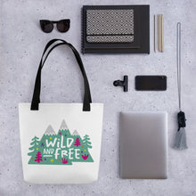 Load image into Gallery viewer, A white tote bag with black handles on a grey tabletop with a laptop and other office supplies. The tote bag has the design with the words &#39;wild and free&#39; with the words inside illustrated mountains with illustrated trees around the words. 