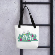 Load image into Gallery viewer, A white tote bag with black handles handing on a black ladder. The tote bag has the design with the words &#39;wild and free&#39; with the words inside illustrated mountains with illustrated trees around the words. 