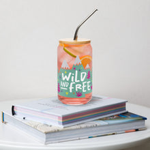 Load image into Gallery viewer, A photo of a glass can tumbler on a coffee table on top of books. The glass tumbler has a bamboo lid and silver metal straw. The design on the glass tumbler says the words &quot;wild and free&quot; inside illustrated mountains with sketchy trees around. 