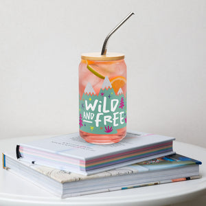 A photo of a glass can tumbler on a coffee table on top of books. The glass tumbler has a bamboo lid and silver metal straw. The design on the glass tumbler says the words "wild and free" inside illustrated mountains with sketchy trees around. 