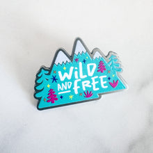 Load image into Gallery viewer, An enamel pin features the words &quot;wild and free&quot; inside illustrated mountains with illustrated trees. 