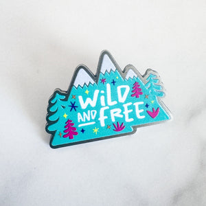 An enamel pin features the words "wild and free" inside illustrated mountains with illustrated trees. 