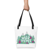 Load image into Gallery viewer, A person holding a white tote bag with black handles. The tote bag has the design with the words &#39;wild and free&#39; with the words inside illustrated mountains with illustrated trees around the words. 