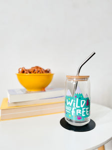 A photo of a glass can tumbler on a coffee table with books and a bowl of pretzels in the background. The glass tumbler has a bamboo lid and silver metal straw. The design on the glass tumbler says the words "wild and free" inside illustrated mountains with sketchy trees around. 