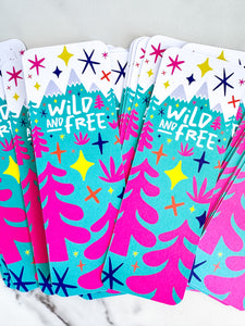 Bookmark placed on a tabletop. The bookmarks feature the words "wild and free" inside illustrated snow-capped mountains with illustrated trees. 