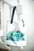 Load image into Gallery viewer, A white tote bag with a black handle hanging on a staircase. The tote bag has the design with the words &#39;wild and free&#39; with the words inside illustrated mountains with illustrated trees around the words. 