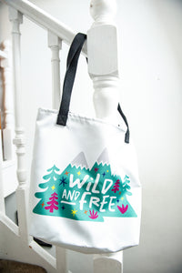 A white tote bag with a black handle hanging on a staircase. The tote bag has the design with the words 'wild and free' with the words inside illustrated mountains with illustrated trees around the words. 