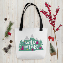 Load image into Gallery viewer, A white tote bag with black handles on a wood tabletop with Christmas items around it. The tote bag has the design with the words &#39;wild and free&#39; with the words inside illustrated mountains with illustrated trees around the words. 