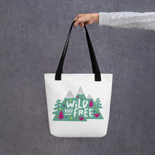 Load image into Gallery viewer, A person holding a white tote bag with black handles. The tote bag has the design with the words &#39;wild and free&#39; with the words inside illustrated mountains with illustrated trees around the words. 