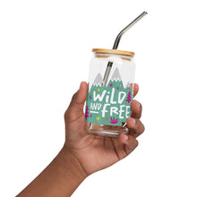 Load image into Gallery viewer, A photo of a glass can tumbler being held in a hand. The glass tumbler has a bamboo lid and silver metal straw. The design on the glass tumbler says the words &quot;wild and free&quot; inside illustrated mountains with sketchy trees around. 