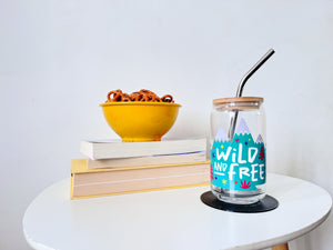 A photo of a glass can tumbler on a coffee table with books and a bowl of pretzels in the background. The glass tumbler has a bamboo lid and silver metal straw. The design on the glass tumbler says the words "wild and free" inside illustrated mountains with sketchy trees around. 