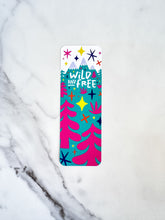 Load image into Gallery viewer, A bookmark placed on a tabletop. The bookmark features the words &quot;wild and free&quot; inside illustrated snow-capped mountains with illustrated trees. 