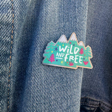 Load image into Gallery viewer, An enamel pin on a jean jacket. The pin features the words &quot;wild and free&quot; inside illustrated mountains with illustrated trees. 